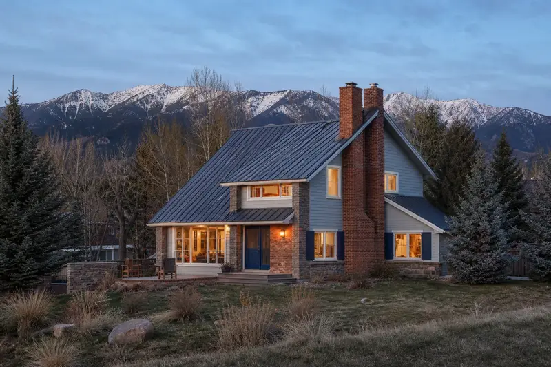 A 1980s mountain modern house with a metal roof with a two-flue brick chimney on a steep gable roof and warm lamp-lit windows; illustrative image
