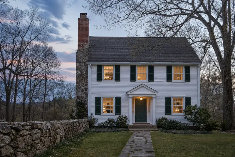 A 1950s colonial with a fieldstone chimney with a tall brick chimney on a steep gable roof of clean red brick; illustrative image