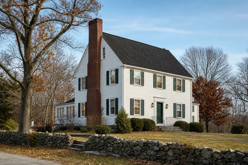 A 1950s white clapboard colonial in soft daylight with a tall brick chimney on a steep gable roof standing clear above the roofline; illustrative image