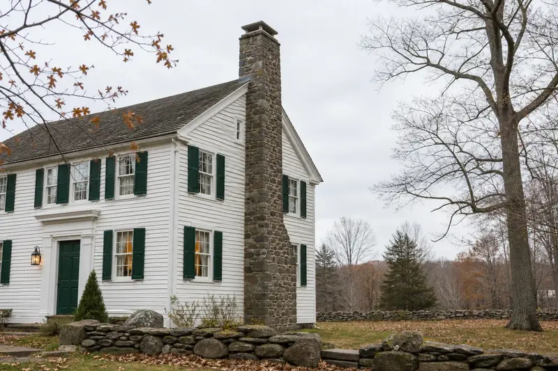 A 1940s white clapboard colonial in soft daylight with a stone masonry chimney standing clear above the roofline; illustrative image