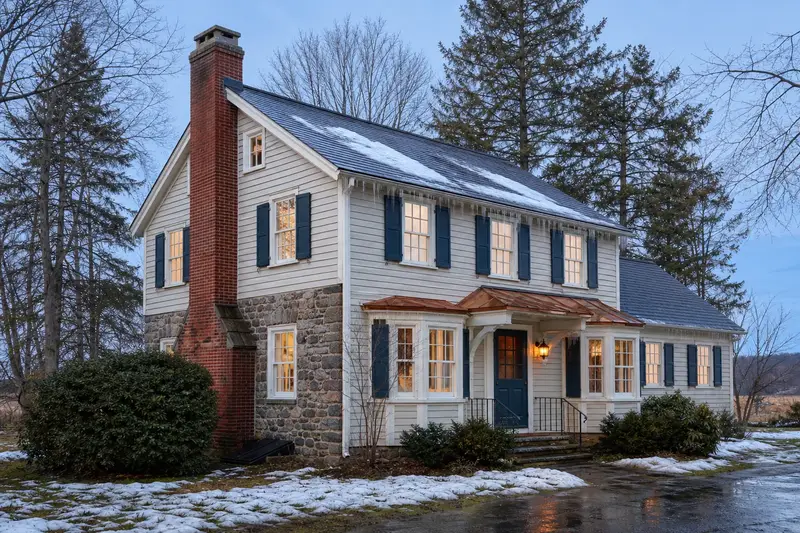 A 1920s stone-and-clapboard farmhouse on a thawing winter day with icicles at the eaves and a red brick chimney with a concrete crown; illustrative image
