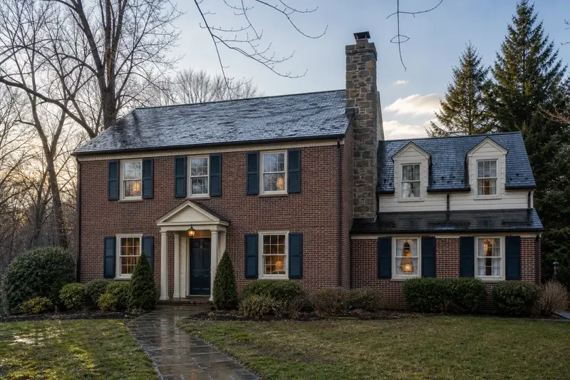 A 1950s brick colonial after a cold rain with a stone masonry chimney, wet roofs and clear gutters; illustrative image