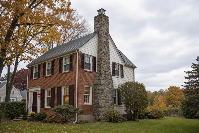 A 1960s brick-front colonial-style suburban house with a fieldstone chimney with a metal cap rising above the roof; illustrative image