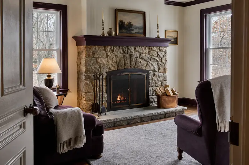 A tidy hearth room with a factory-built wood-burning fireplace with a stone facing, a cleared hearth, fireplace tools and split firewood; illustrative image