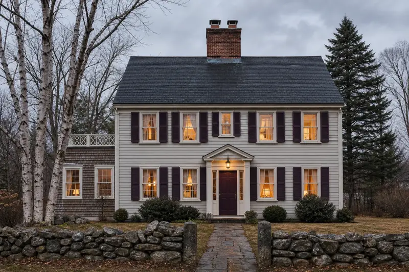 A 1950s clapboard colonial with a two-flue brick chimney on a slate roof and warm lamp-lit windows; illustrative image