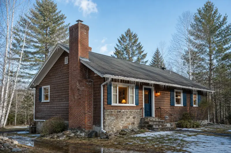 A 1950s cedar-sided rambler in early thaw with icicles at the eaves and a tall brick chimney on a steep gable roof; illustrative image