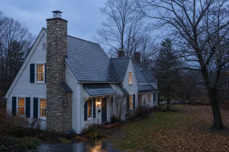 A 1940s steep-gabled frame house after steady rain with a stone masonry chimney and a stainless steel cap; illustrative image