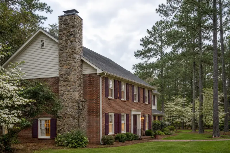 A 1990s two-story brick colonial-style suburban house in soft daylight with a fieldstone chimney with a metal cap standing clear above the roofline; illustrative image
