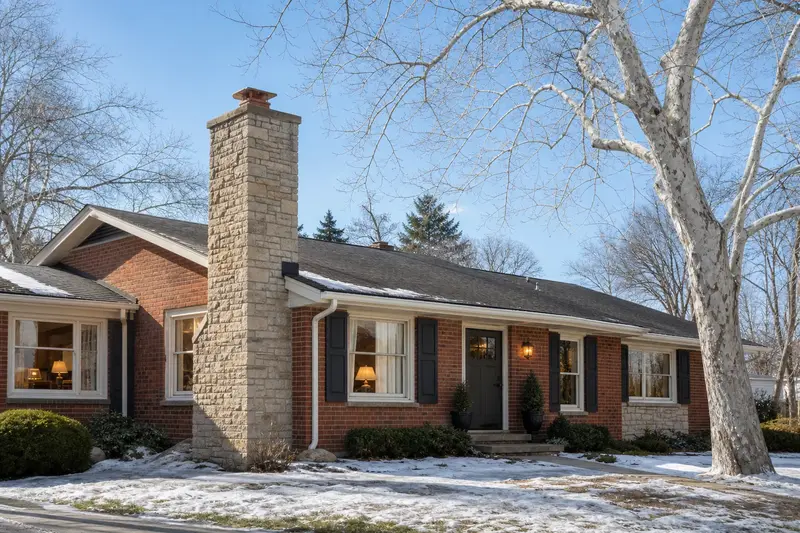 A 1960s brick-front ranch house with a stone masonry chimney and warm windows as the heating season begins; illustrative image