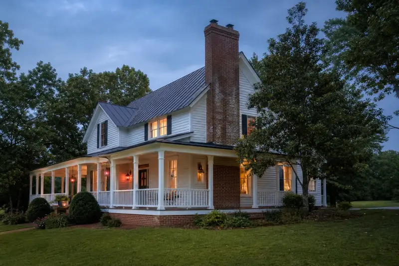 A 1980s farmhouse-style house with a wraparound porch with a two-flue brick chimney on a steep metal roof of clean red brick; illustrative image