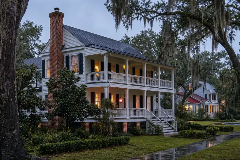 A 1940s raised Lowcountry-style house with a deep two-story porch after a heavy rain with a brick chimney with a stainless steel cap on an asphalt shingle roof, wet roofs and clear gutters; illustrative image