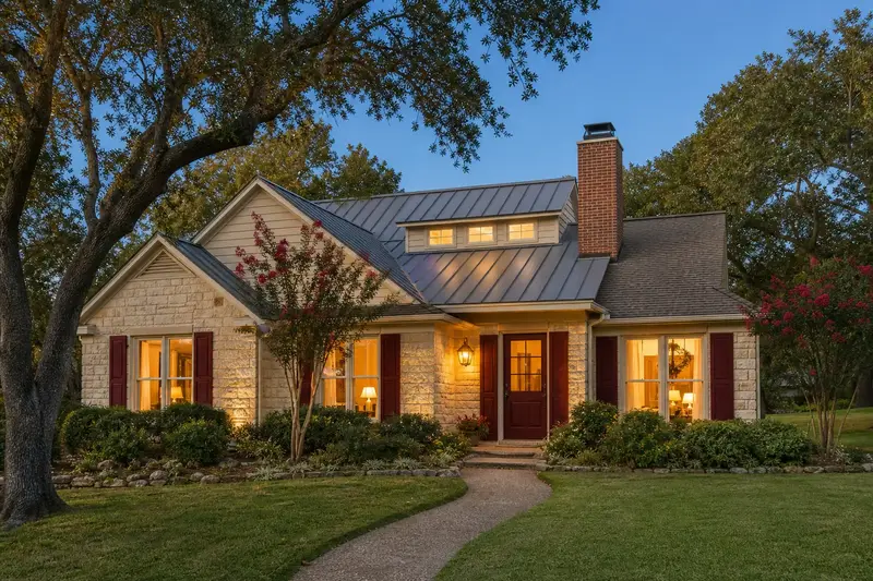 A 1980s limestone contemporary house with a metal roof with a brick chimney with a metal cap on an asphalt shingle roof and warm lamp-lit windows; illustrative image