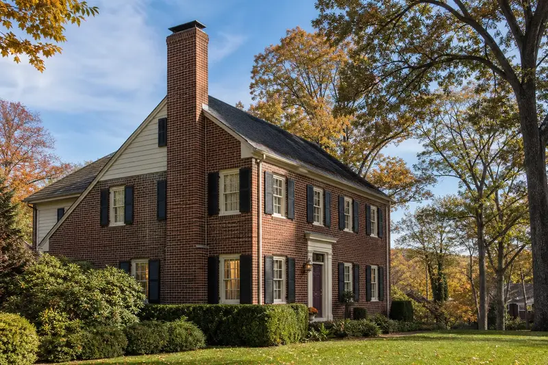 A 1950s brick colonial with black shutters in soft daylight with an exterior end brick chimney on a gable roof standing clear above the roofline; illustrative image